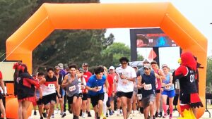 Runners take off from the starting line under a large orange inflatable arch at the beginning of a 5K race. Participants wear numbered bibs and athletic gear, with a mix of serious and excited expressions. On either side of the arch, Howard College cheerleaders and the school mascot, a red hawk in a black jersey labeled '01,' cheer on the runners. A digital scoreboard is visible in the background, and trees line the course.